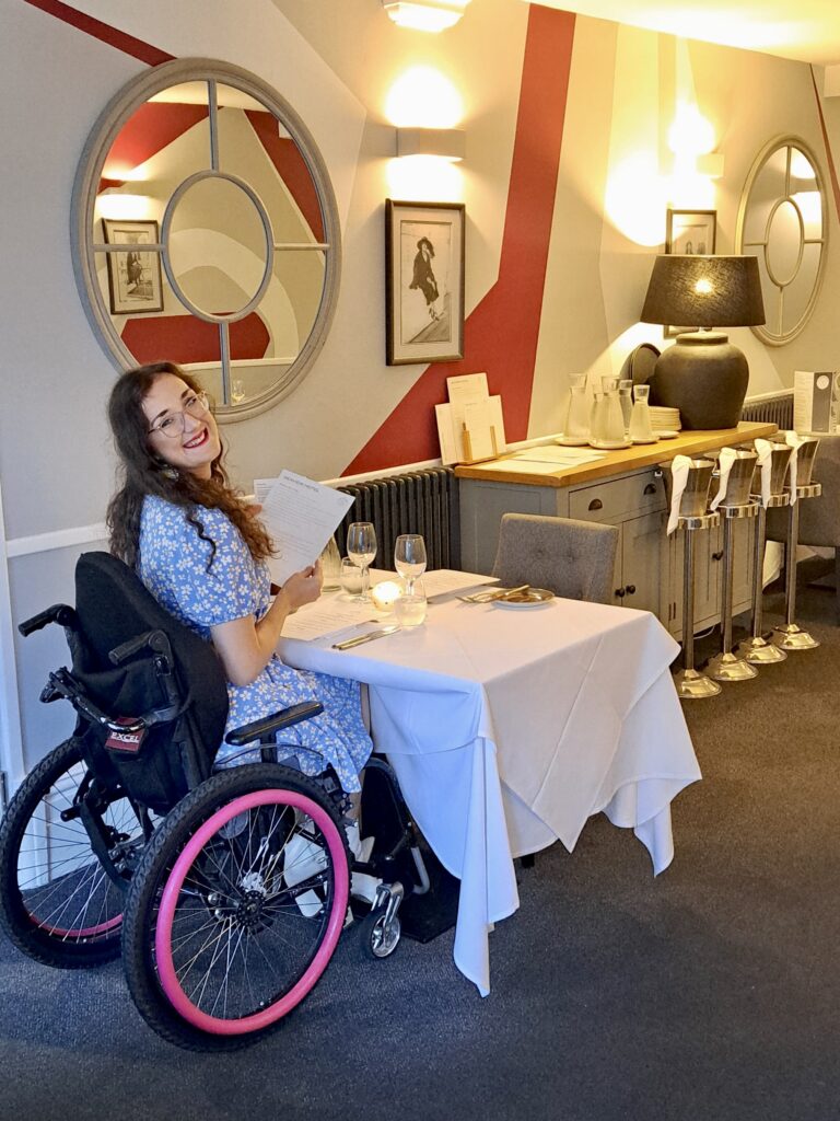 Georgina, a white brunette female who uses a wheelchair, looks back and smiles at the camera in the dining area of The Seaview Hotel in the Isle of Wight